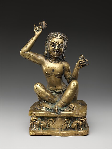 A golden statue of a man. They are shirtless, sitting cross-legged, with a mustache, arms raised overhead holding cymbals.